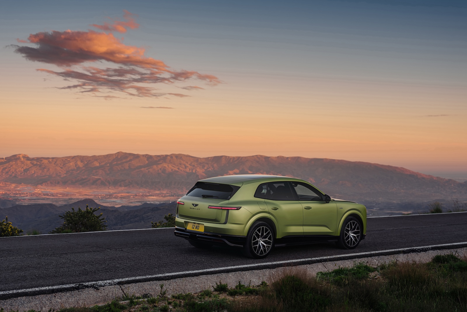 Zielony SUV marki Bentley zaparkowany na górskiej drodze o zachodzie słońca, pośród chmur.