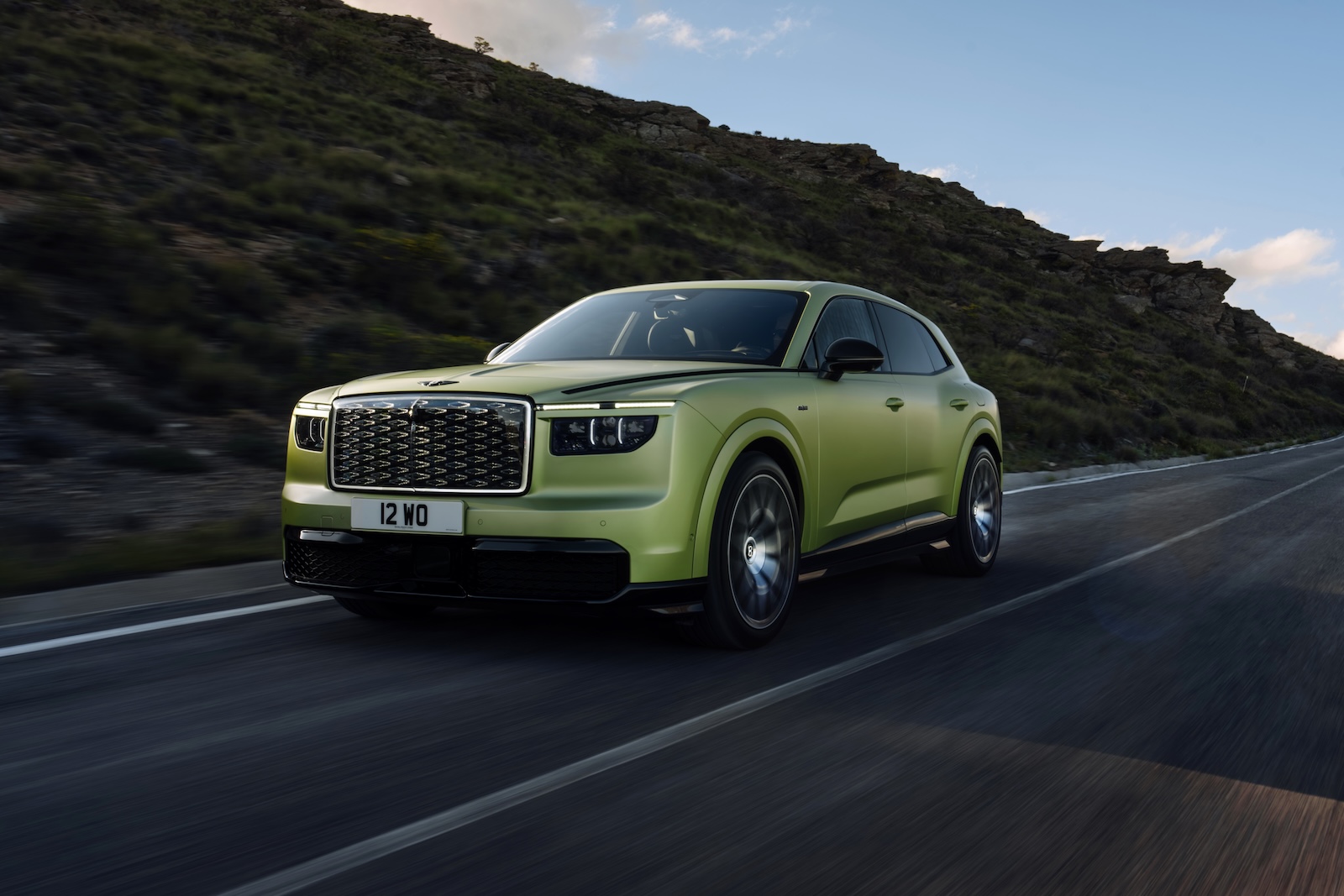 Zielony SUV marki Bentley jedzie górską drogą, a w tle widać wzgórza i chmury.