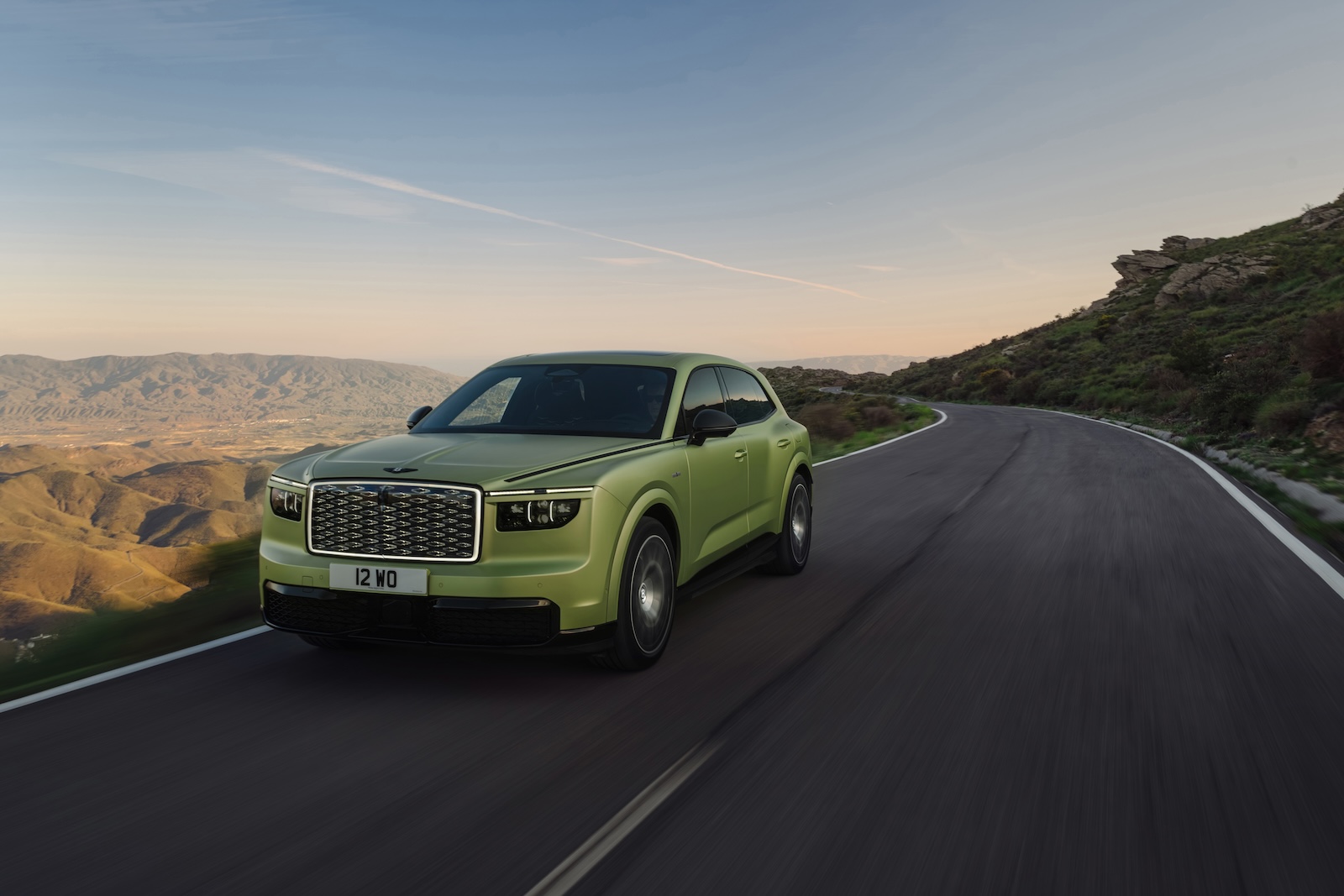 Zielony SUV marki Bentley jedzie krętą górską drogą o zachodzie słońca, okrążając wzgórze.