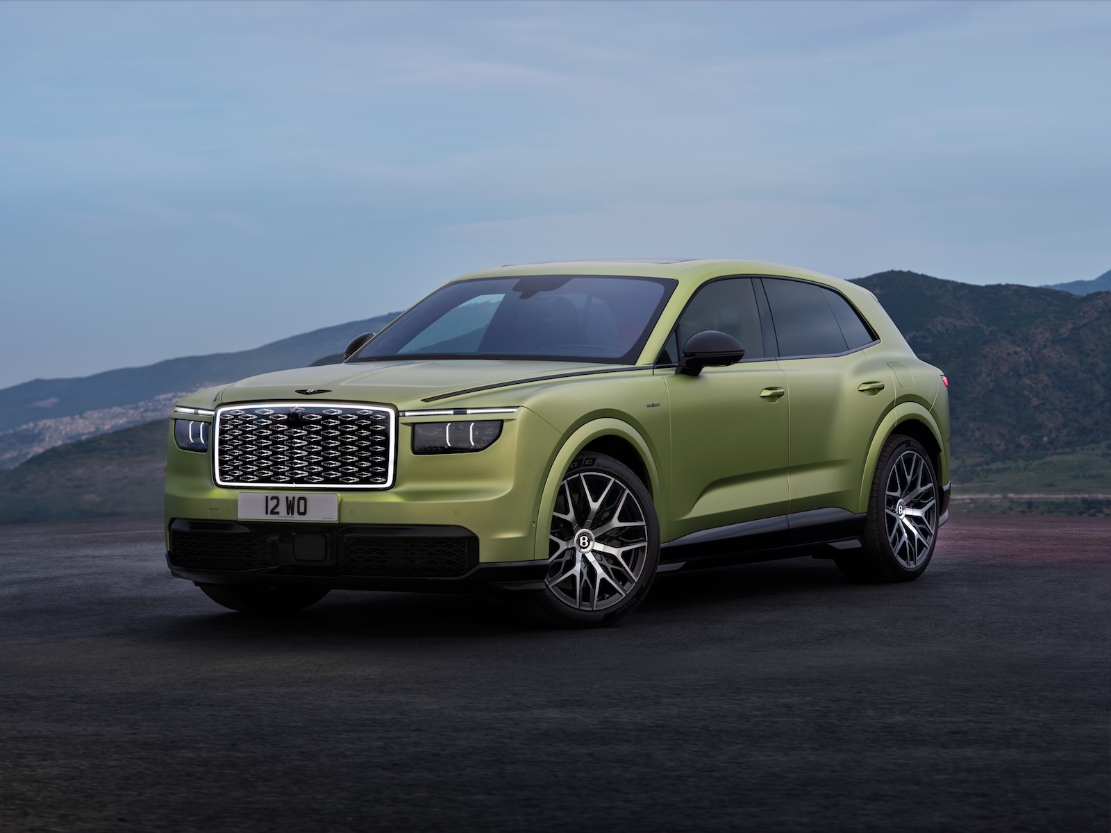 Zielony SUV marki Bentley z czarnymi felgami stoi na asfalcie, a w tle widać góry i chmury.