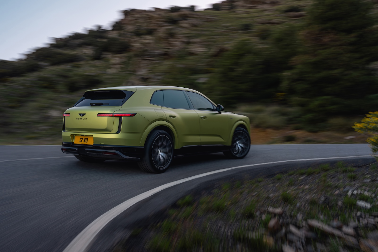 Zielony SUV marki Bentley jedzie krętą górską drogą, otoczony zamazanymi drzewami i skałami.