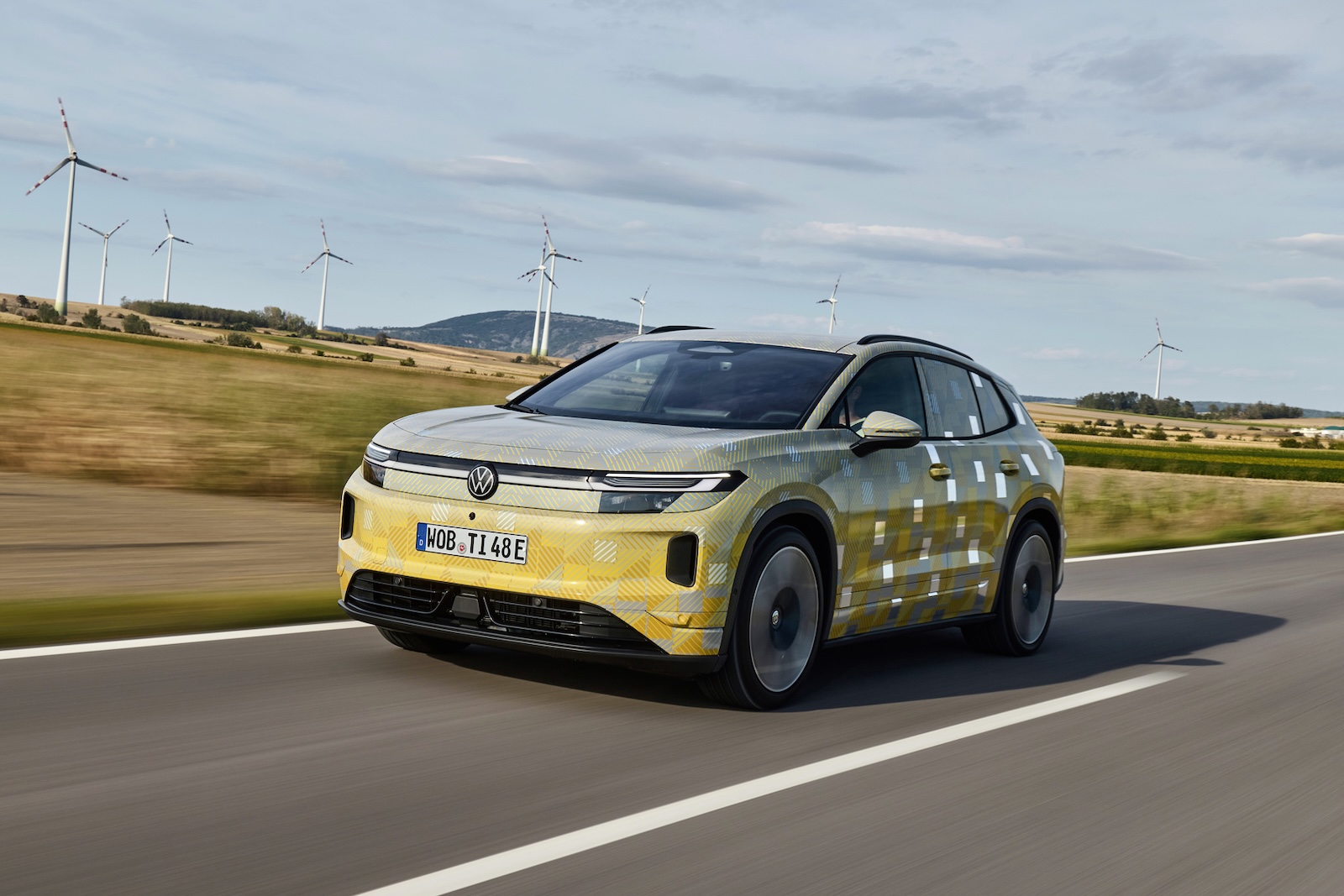 Volkswagen ID. – elektryczny SUV na wiejskiej drodze, wiatraki i pola, częściowo zachmurzone niebo.