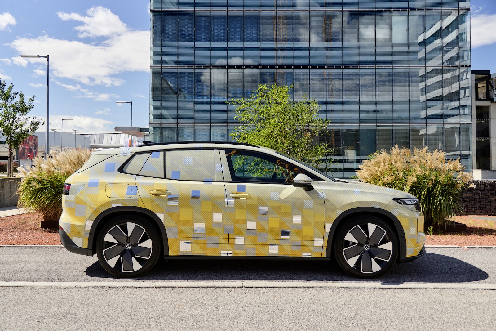 Żółto-niebieski Volkswagen ID. Tiguan stoi przed szklanym biurowcem, otoczony trawą i roślinami.
