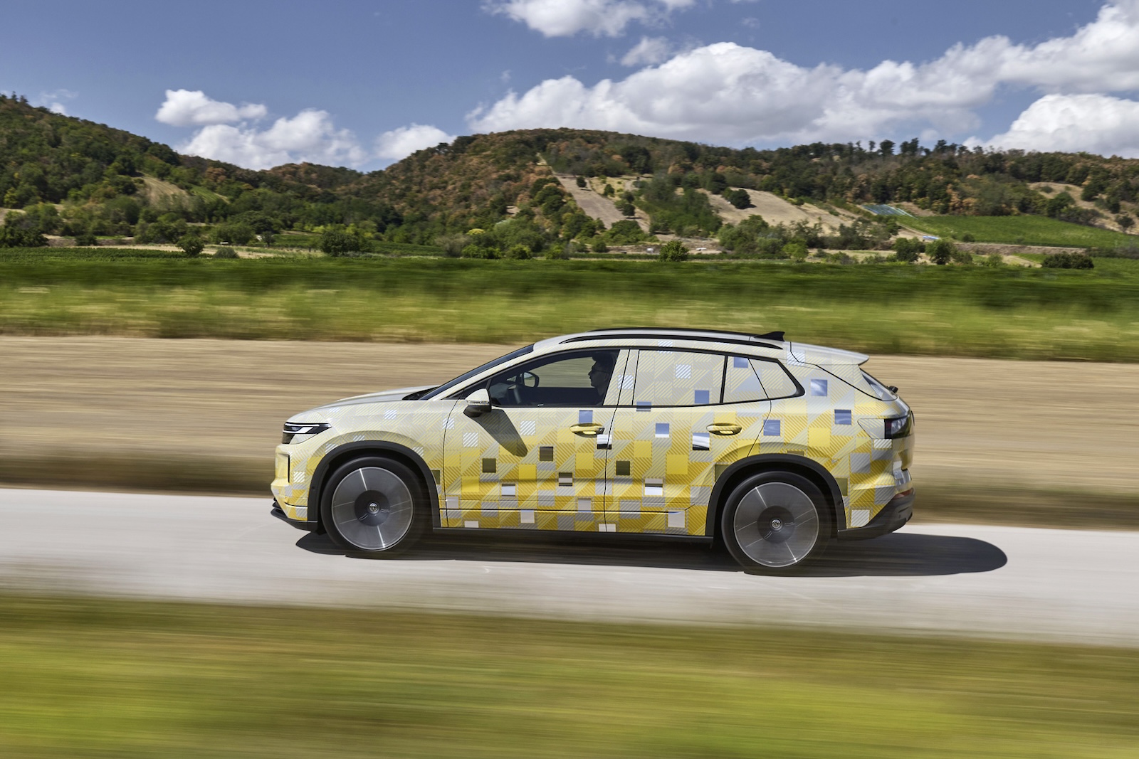 Zakamuflowany prototyp. SUV Tiguan jedzie po asfaltowej drodze wśród zielonych wzgórz i drzew.