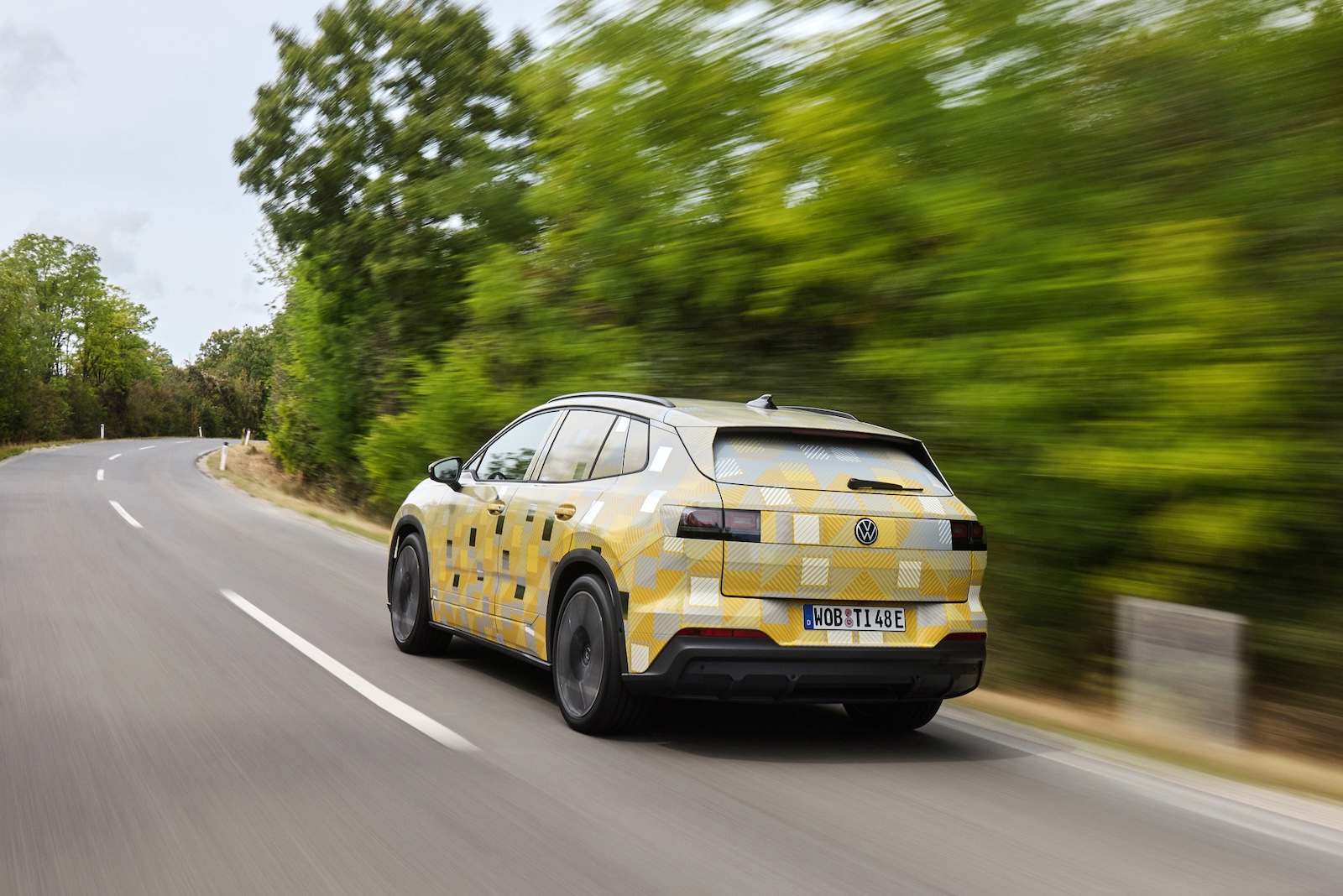 Żółto-szary Volkswagen ID. Tiguan jedzie krętą drogą wśród zielonych drzew, oddalając się.