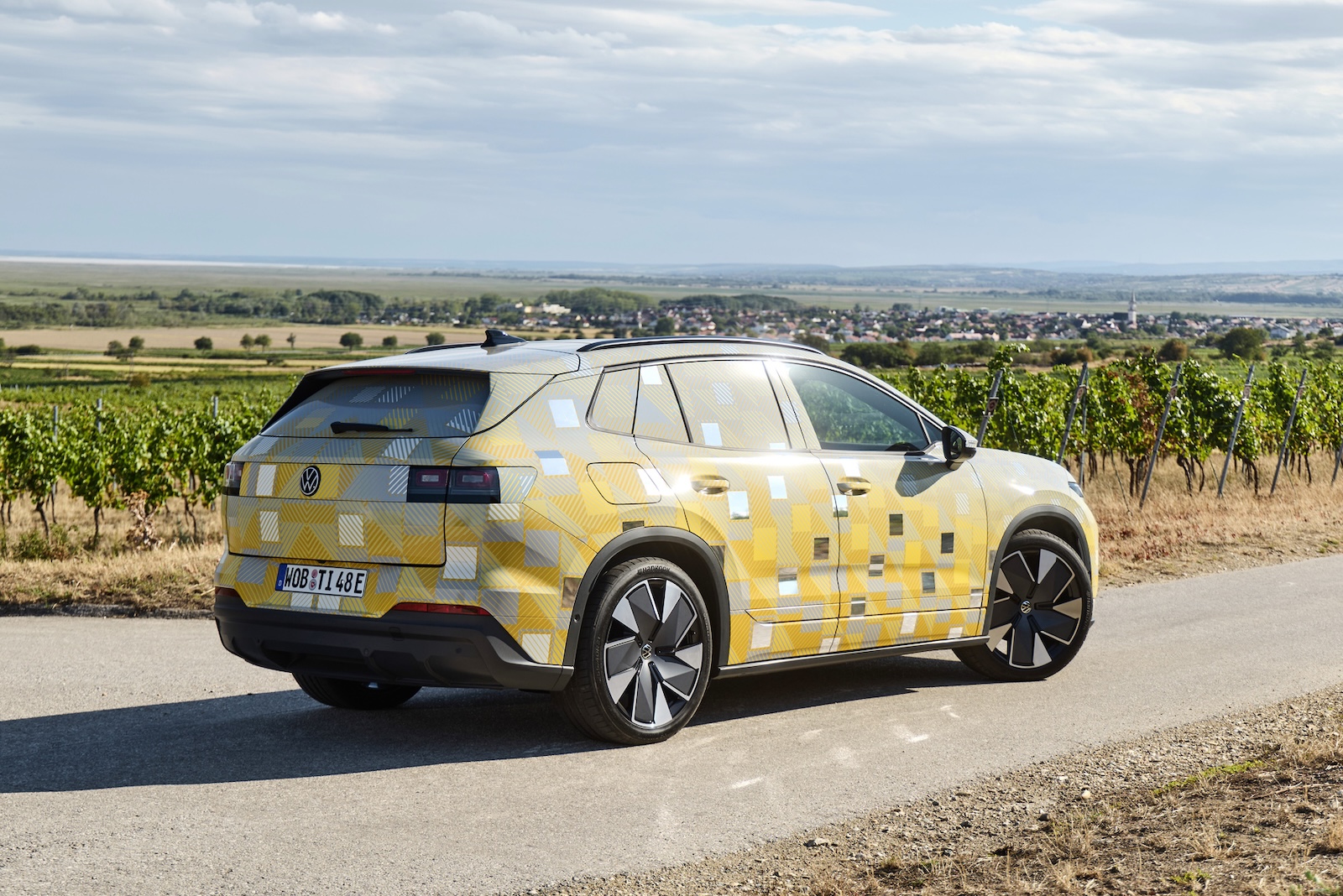 Żółto-szary, zamaskowany SUV ID. Tiguan stoi na wiejskiej drodze obok winnic i pól.