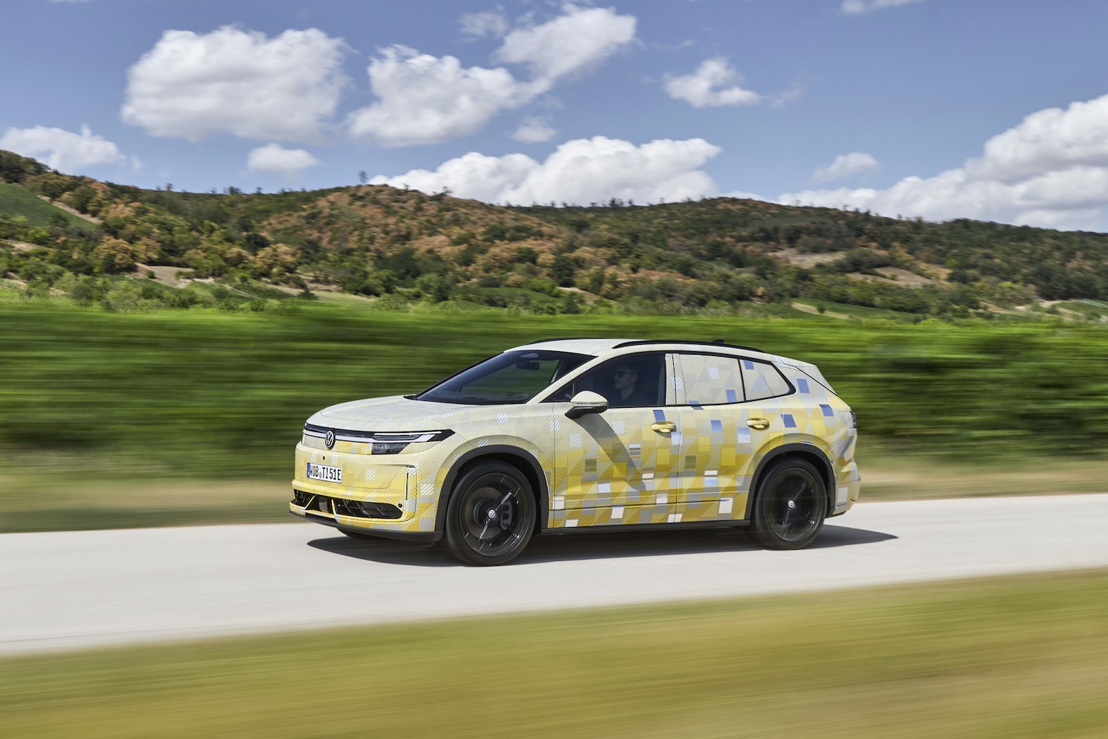 Zakamuflowany model Tiguan jedzie wiejską drogą, mijając zielone pola i wzgórza.