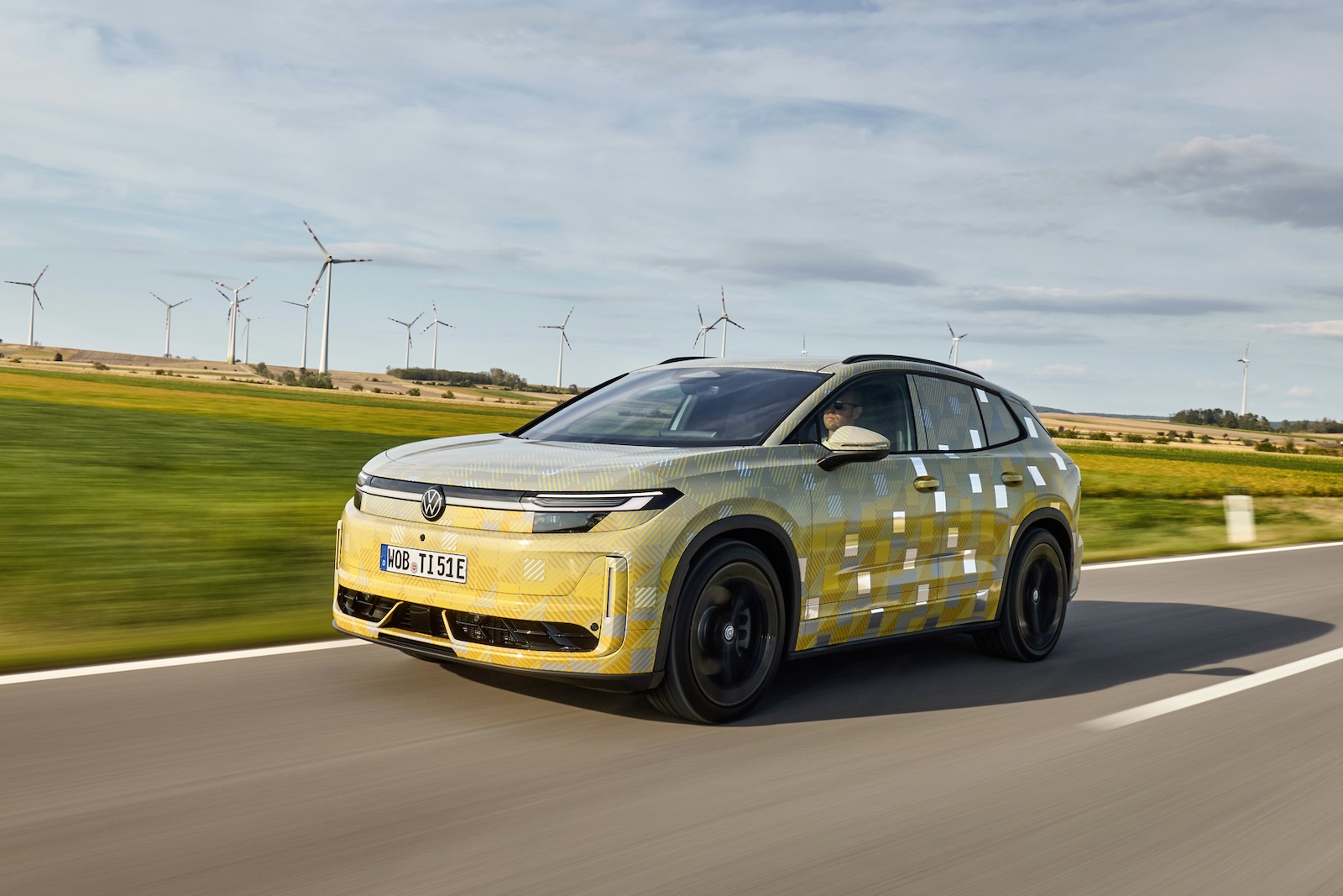 Żółty, zamaskowany Volkswagen ID. Tiguan jedzie wiejską drogą, a w tle widać wiatraki i pola.