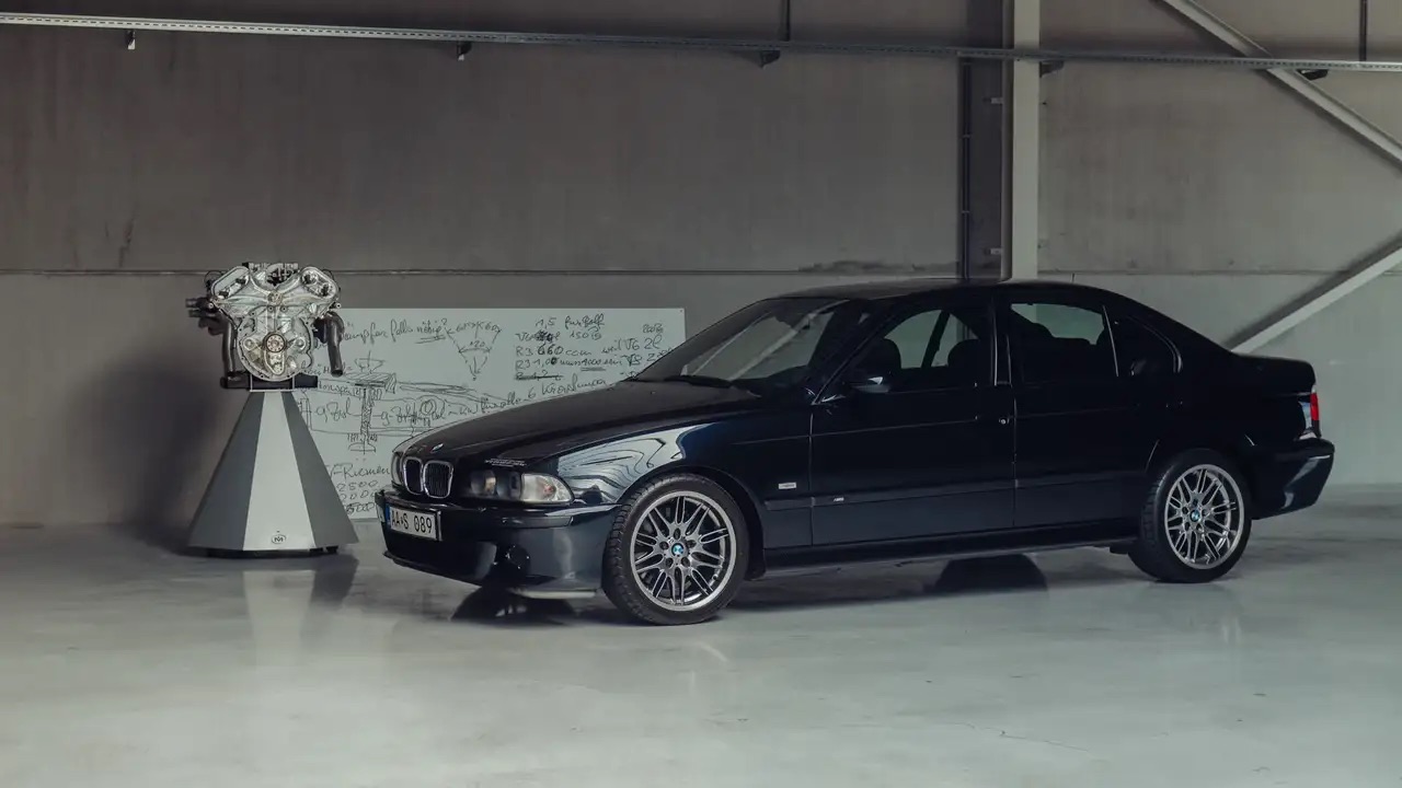 Czarny sedan BMW stoi na białej podłodze obok silnika i tablicy z notatkami.