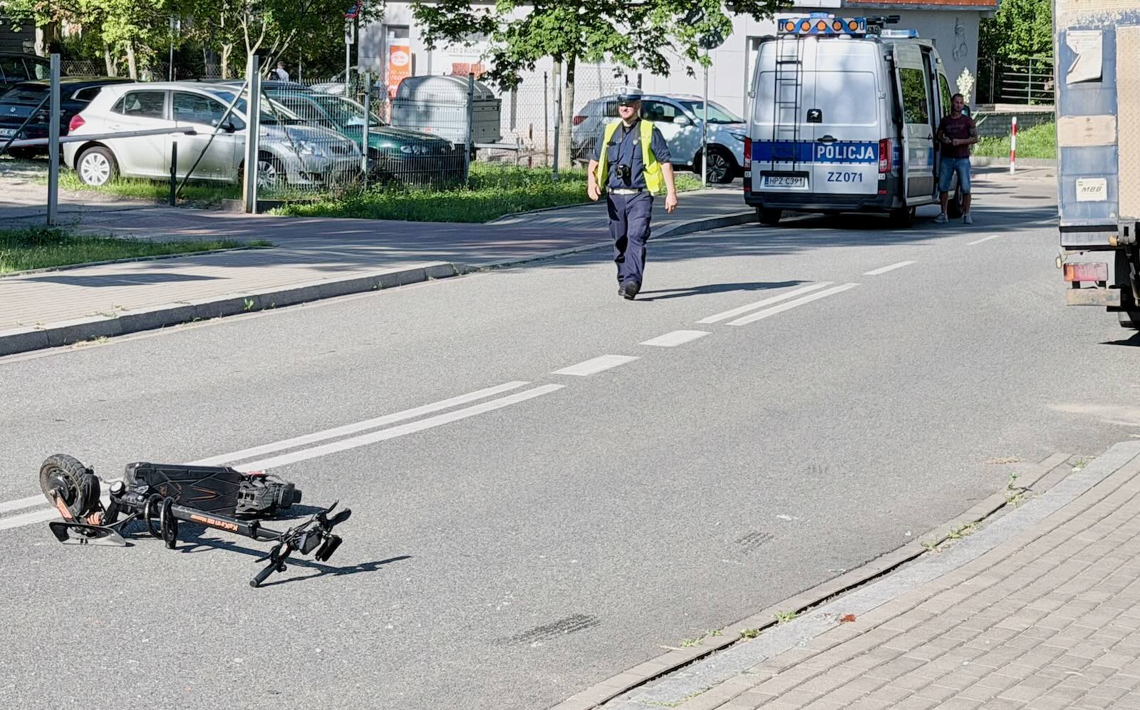 Policjant stoi przy przewróconej hulajnodze na ulicy; w tle widać radiowóz i drugą osobę.