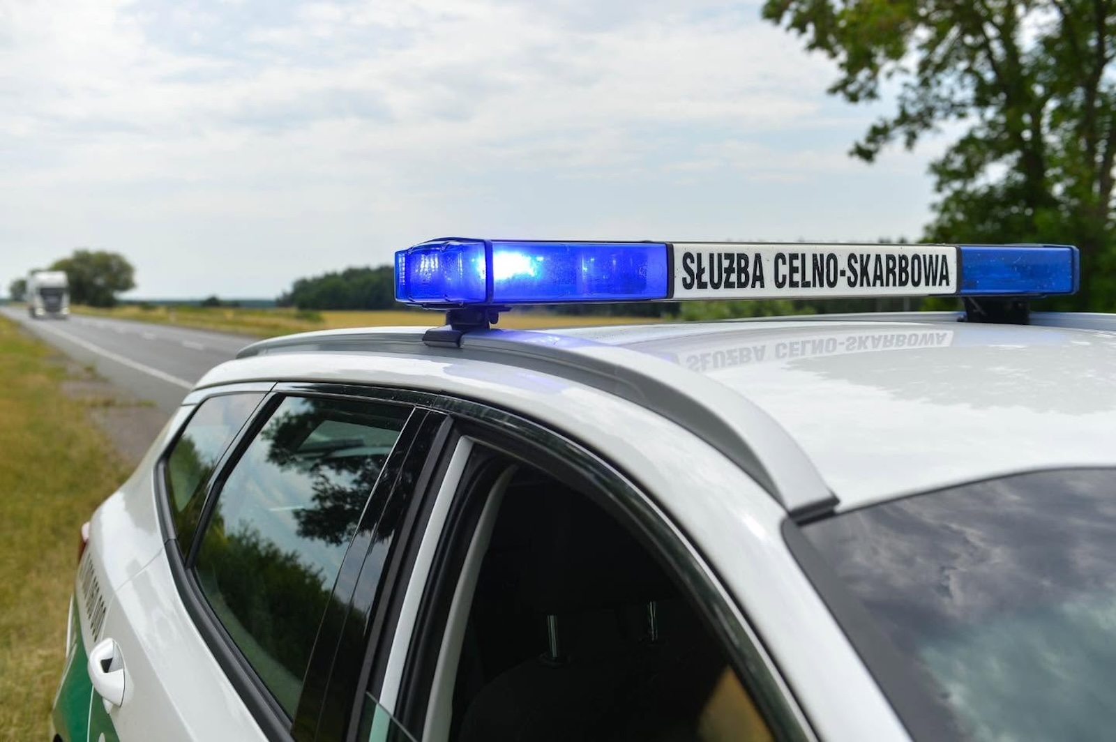 Polski radiowóz służby celno-skarbowej z niebieskimi światłami stoi przy wiejskiej drodze, a w oddali widać ciężarówkę.