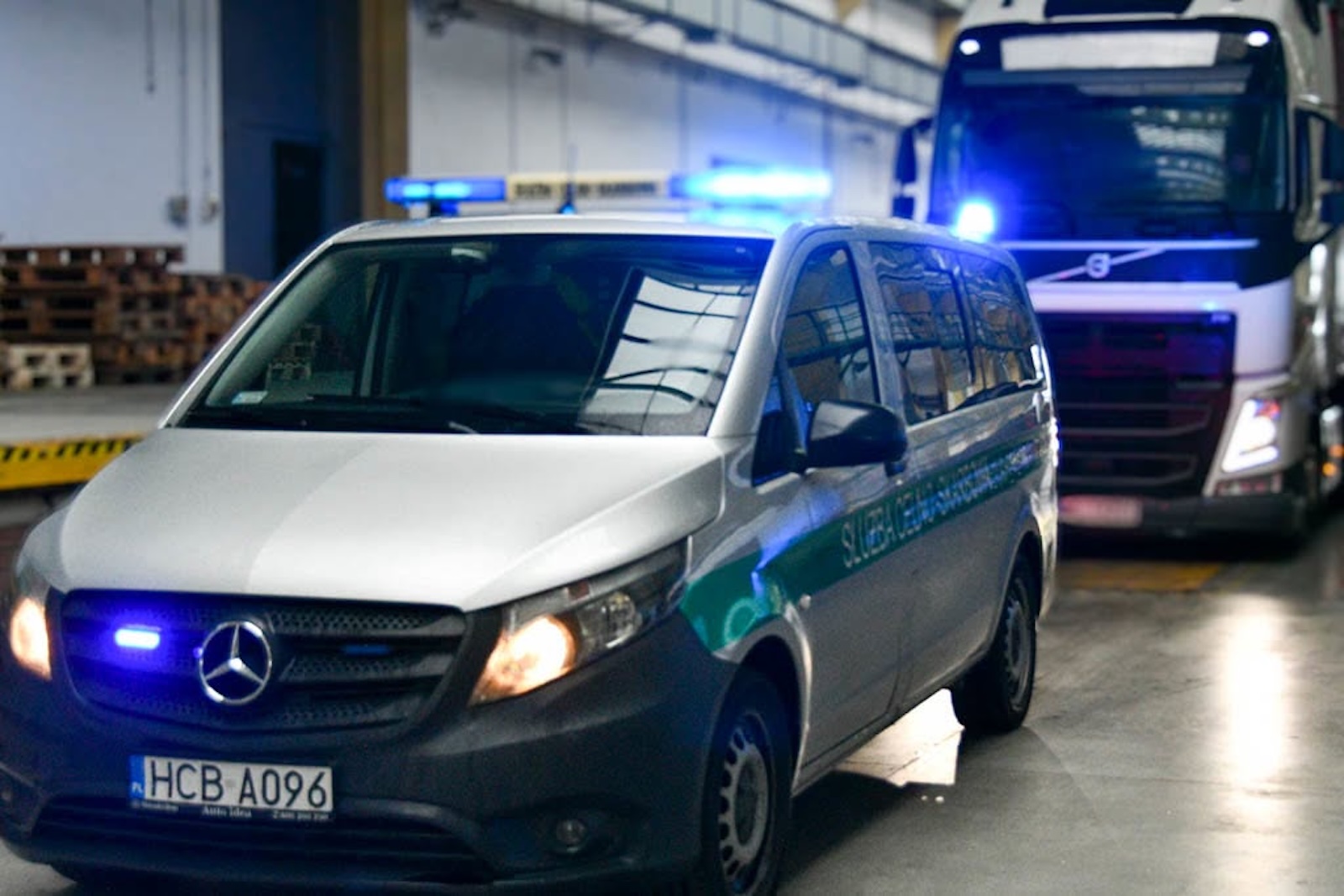 Mercedes z niebieskimi światłami stoi przed białą ciężarówką Volvo wewnątrz budynku.