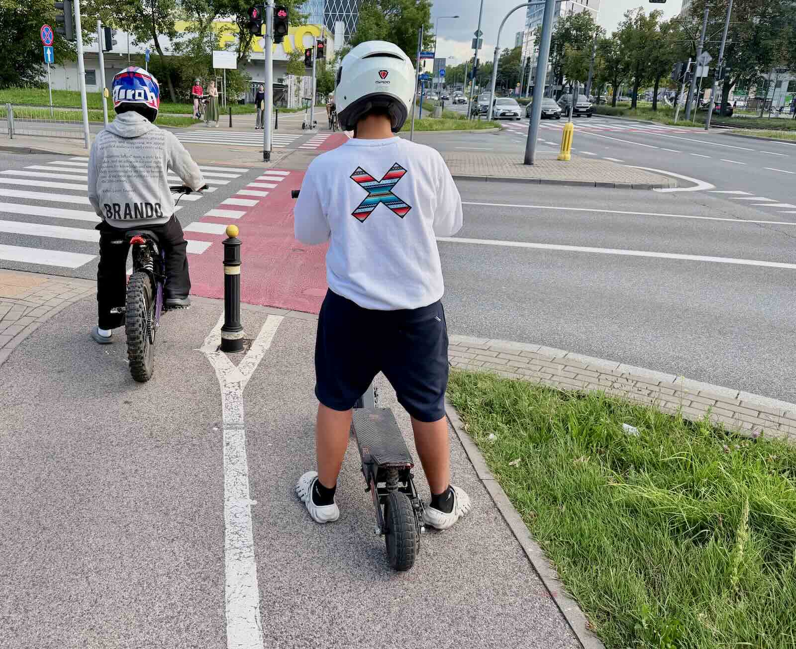 Dwie osoby w kaskach przy przejściu dla pieszych, jedna na rowerze, druga na hulajnodze elektrycznej.