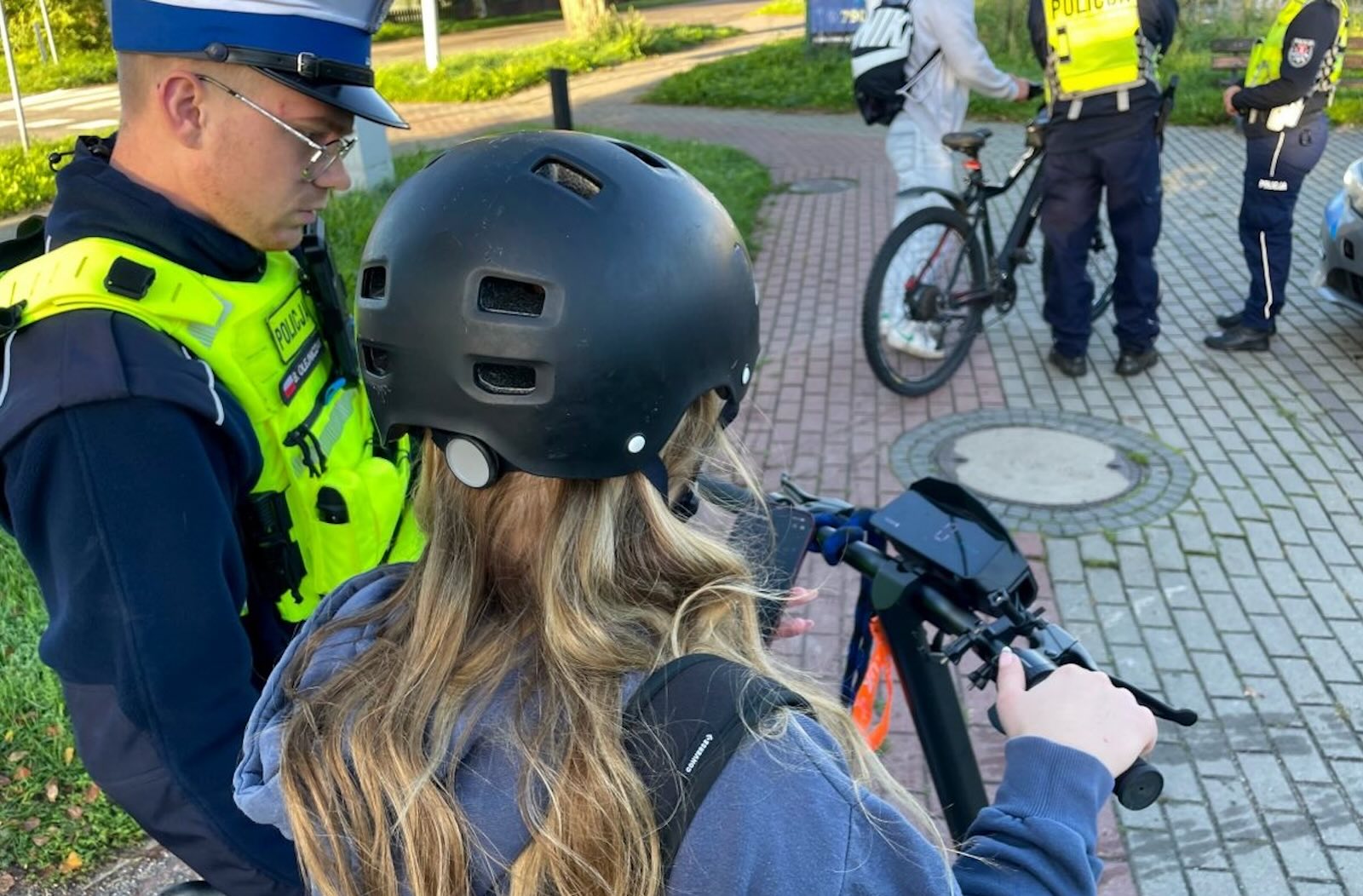 Policjant rozmawia z osobą w kasku stojącą przy hulajnodze; w tle widać dwóch policjantów i rowerzystę.