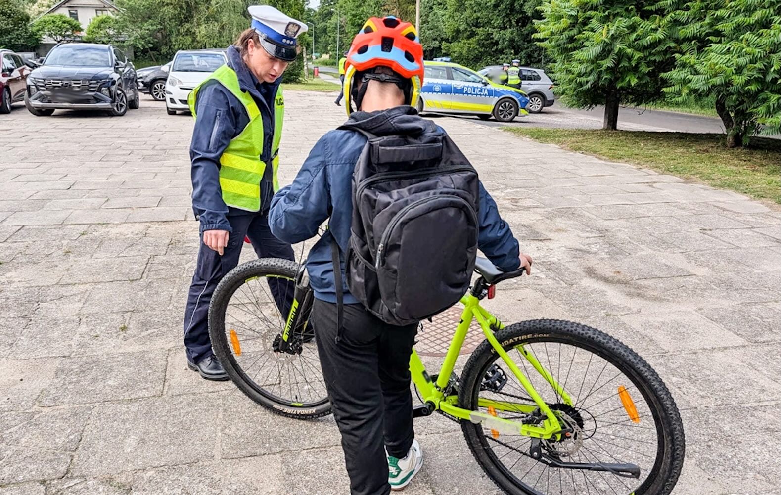 Policjant sprawdza rower, rozmawiając z osobą w kasku na parkingu, na którym stoją samochody i rosną drzewa.