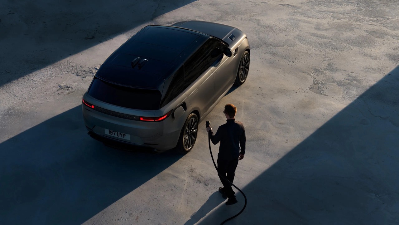 Osoba stojąca przy elektrycznym SUV-ie podłącza kabel do ładowania; na betonie rzucają się długie cienie.