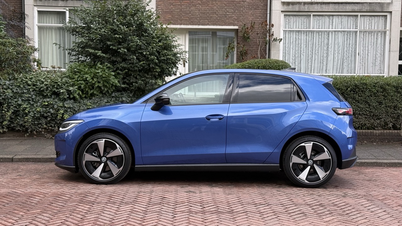 Niebieski hatchback zaparkowany na brukowanej ulicy przed domami porośniętymi krzewami i z oknami.