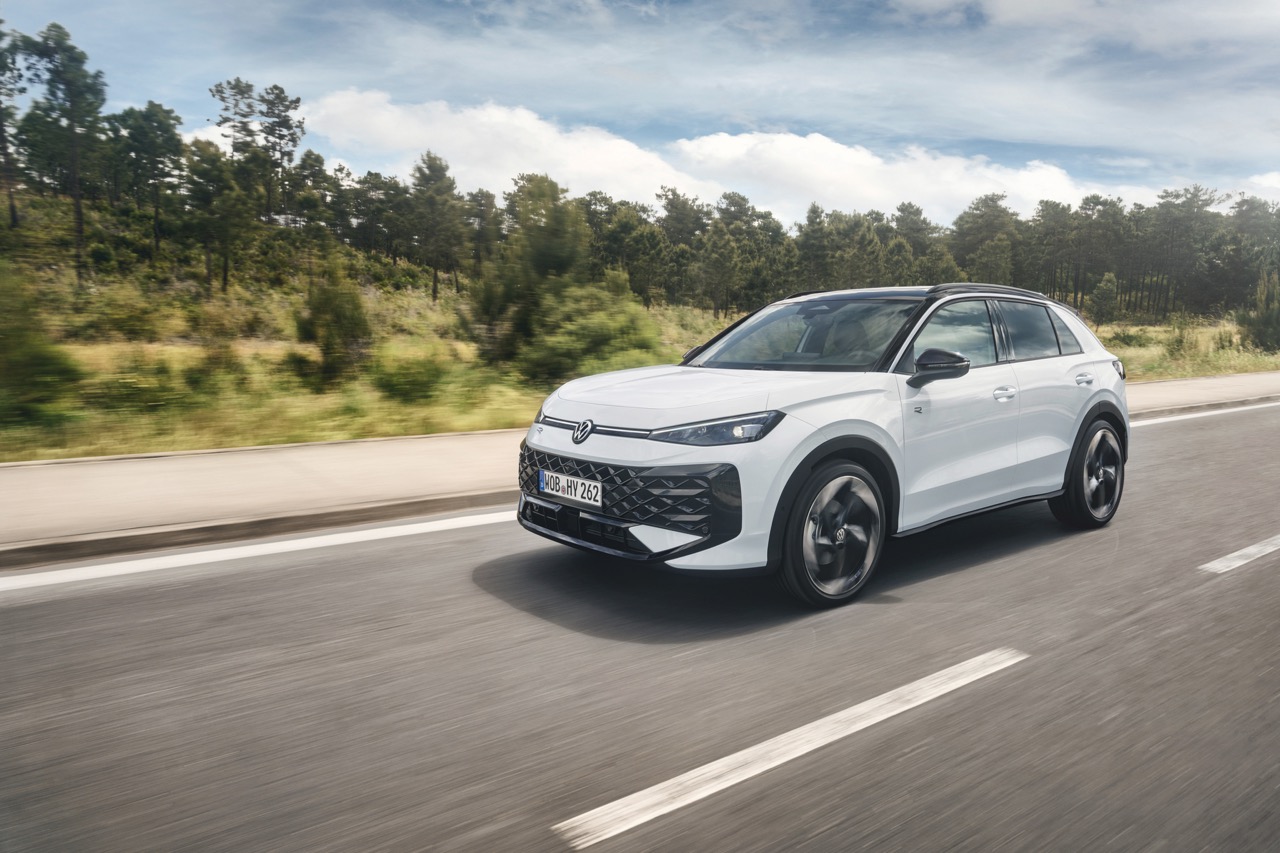 Biały SUV marki Volkswagen jedzie po asfaltowej drodze wzdłuż zieleni i drzew pod chmurami.