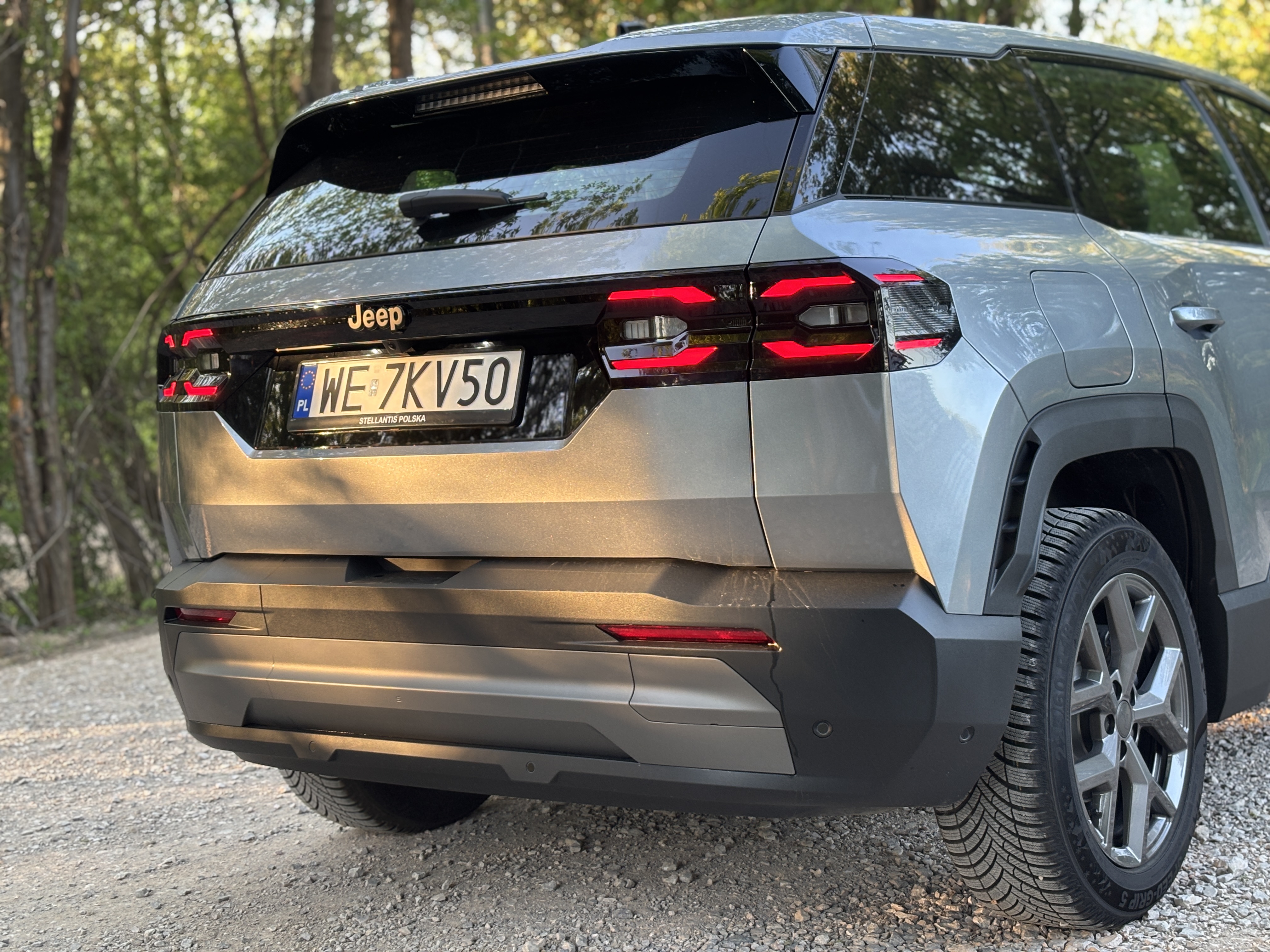 Jasnoszary SUV marki Jeep zaparkowany tyłem na żwirowej ścieżce.