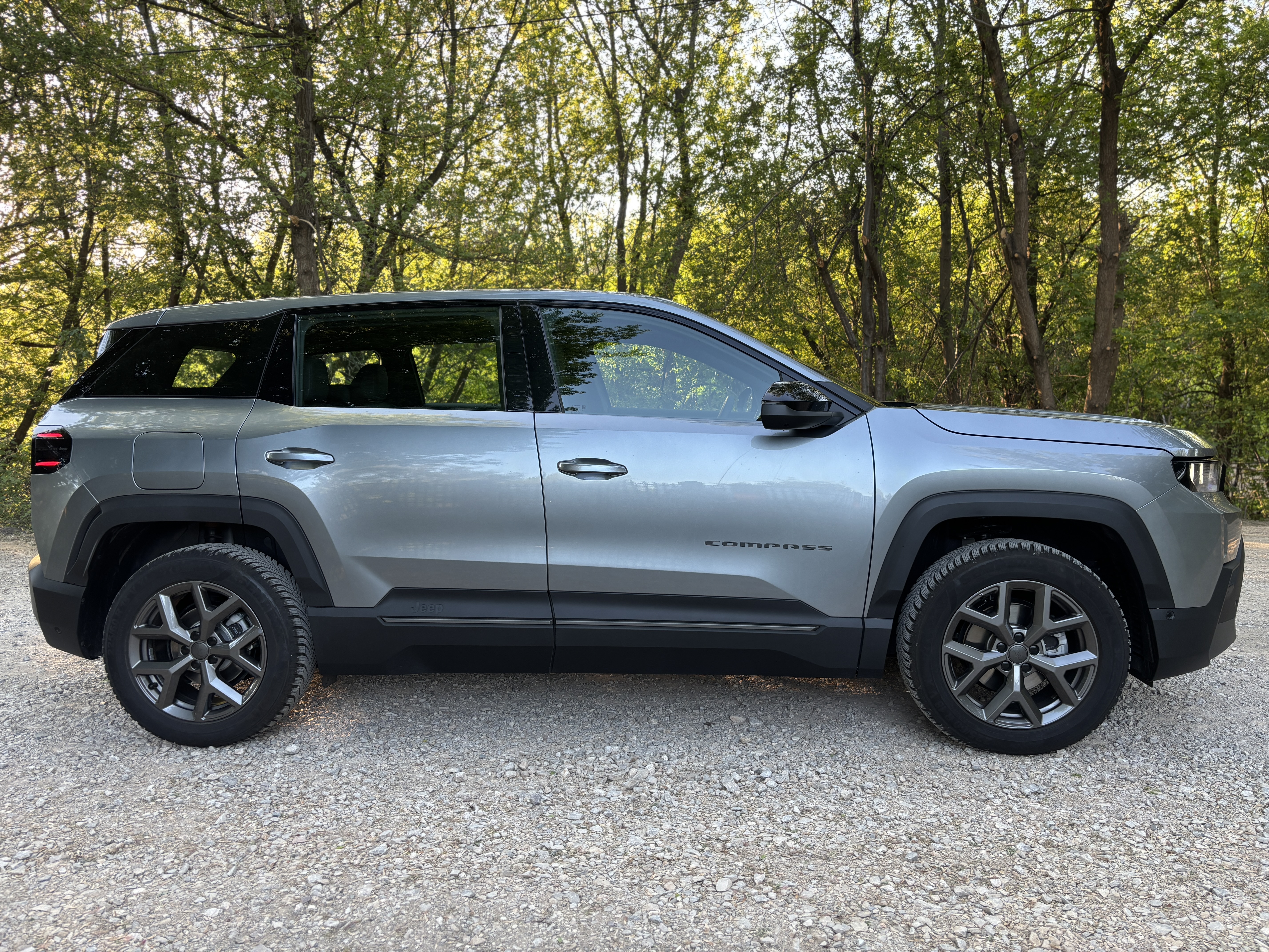 Srebrny Jeep Compass zaparkowany bokiem na żwirowej ścieżce na tle drzew.