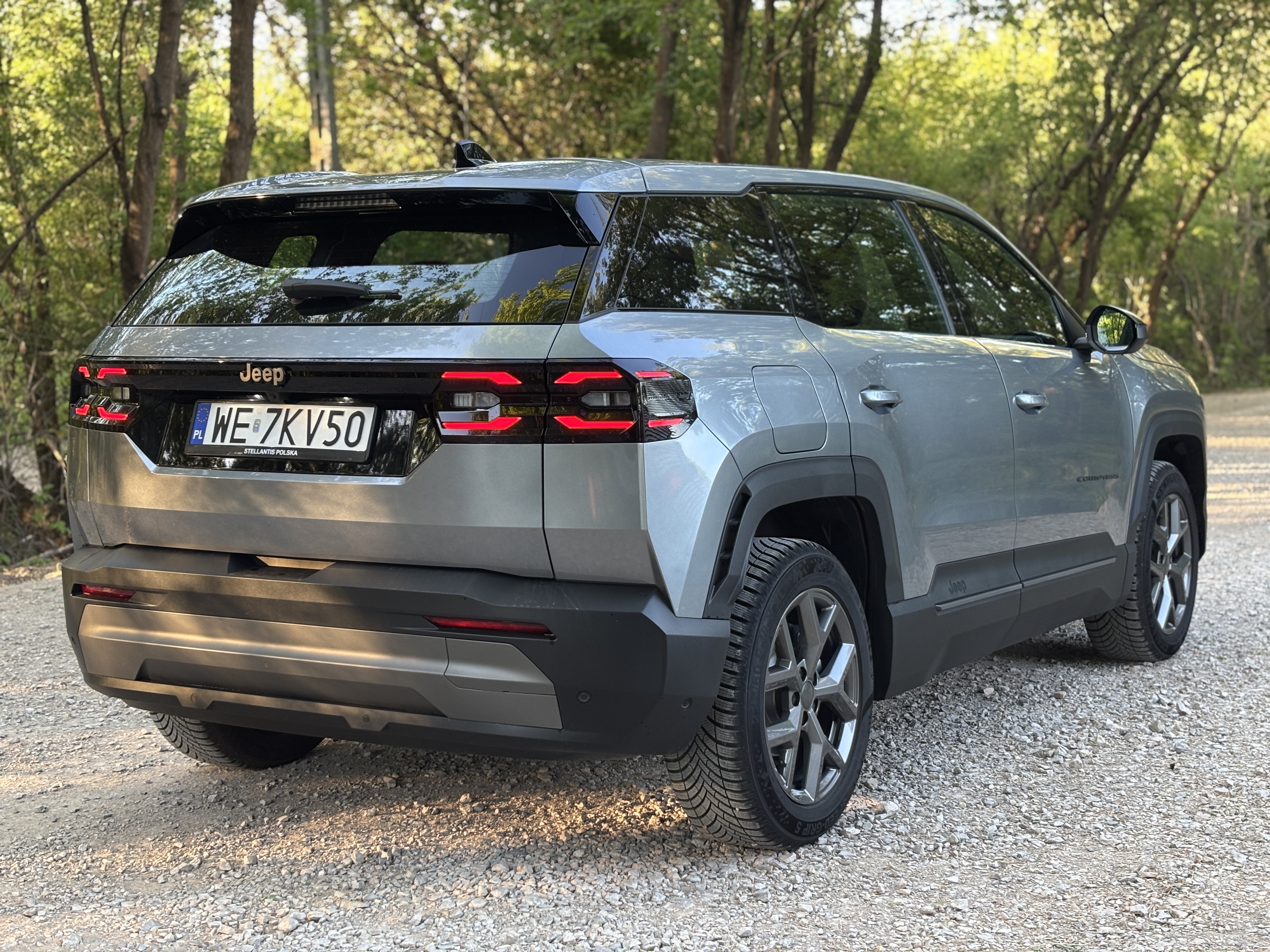 Szary Jeep Compass zaparkowany na żwirowej ścieżce w lesie.