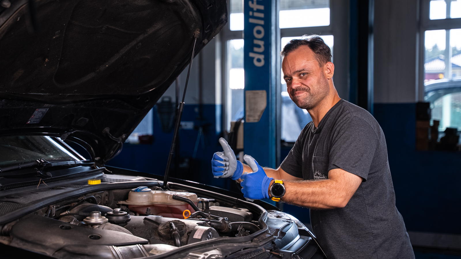 Mechanik w niebieskich rękawiczkach pokazuje kciuk w górę przy silniku używanego VW Passata B5.