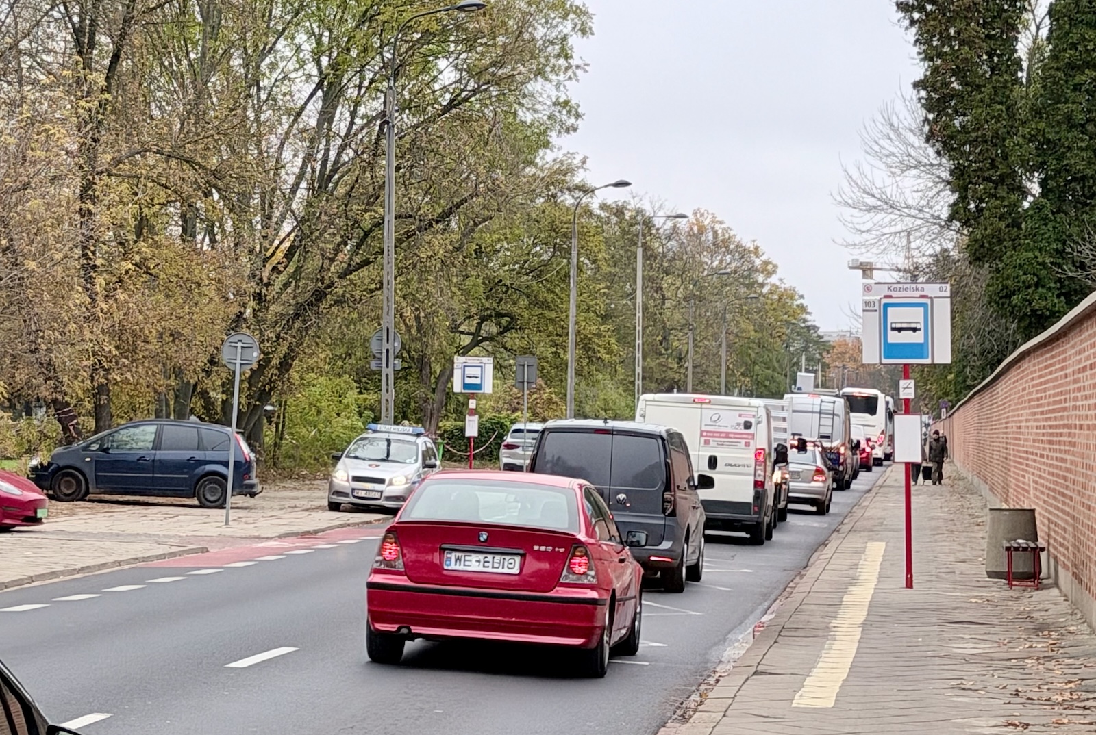 Czerwony samochód w korku przy przystanku autobusowym na miejskiej ulicy obok cmentarza.