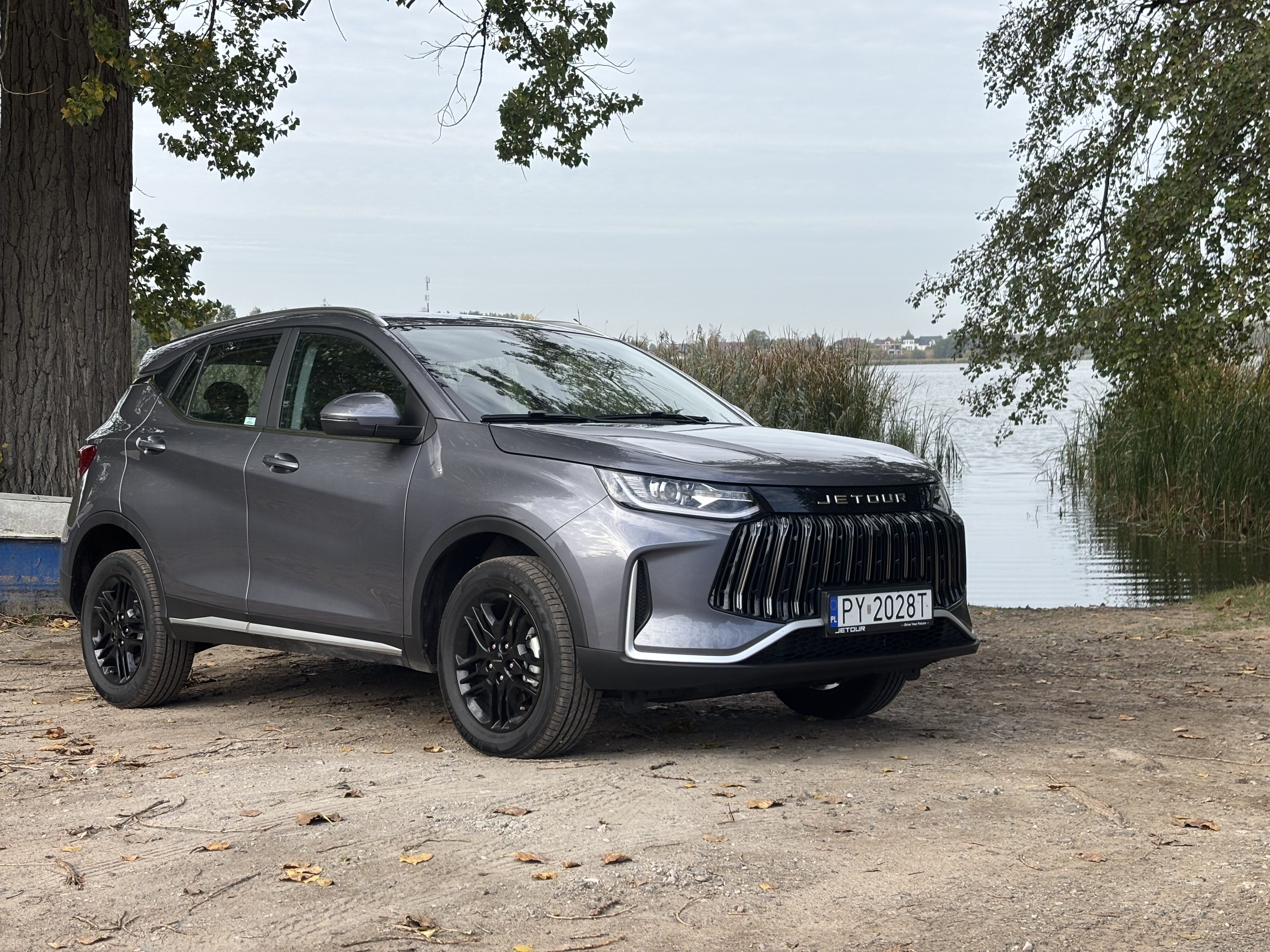 Szary SUV marki Jetour zaparkowany na polnej drodze nad jeziorem.