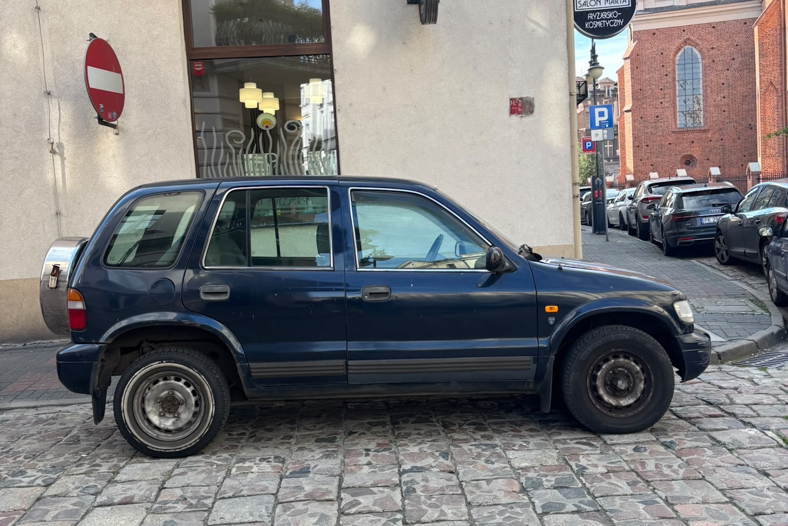 Ciemnoniebieski SUV marki Kia zaparkowany na brukowanej ulicy przy budynku.