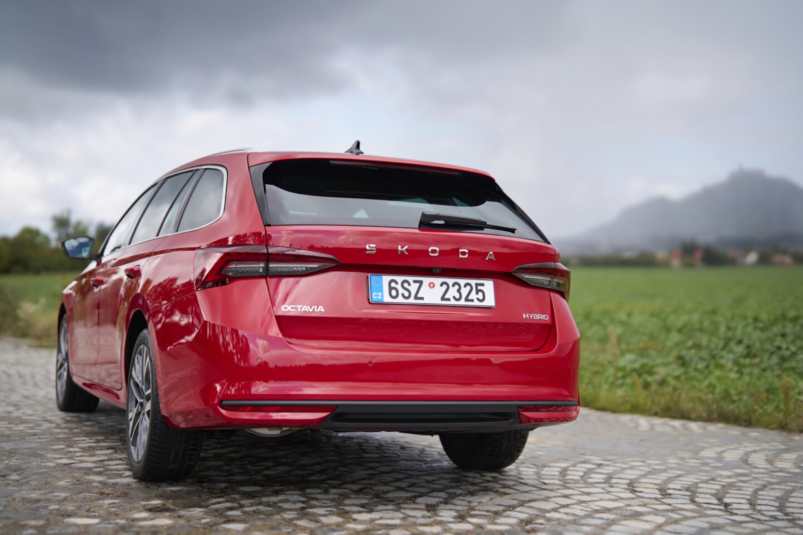 Czerwona Skoda Octavia Hybrid na brukowanej drodze obok zielonego pola, a w tle wzniesienia.