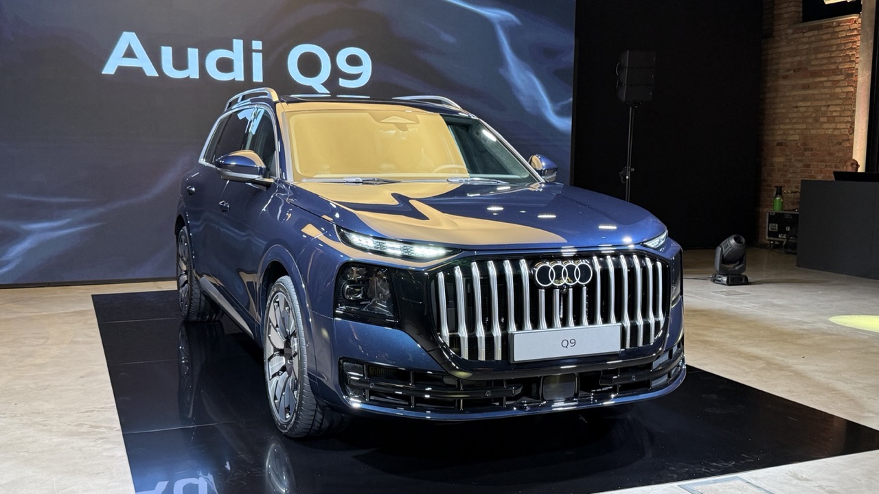 Niebieski SUV Audi Q9 stoi na czarnej platformie, a w tle widać ekran z napisem Audi Q9.