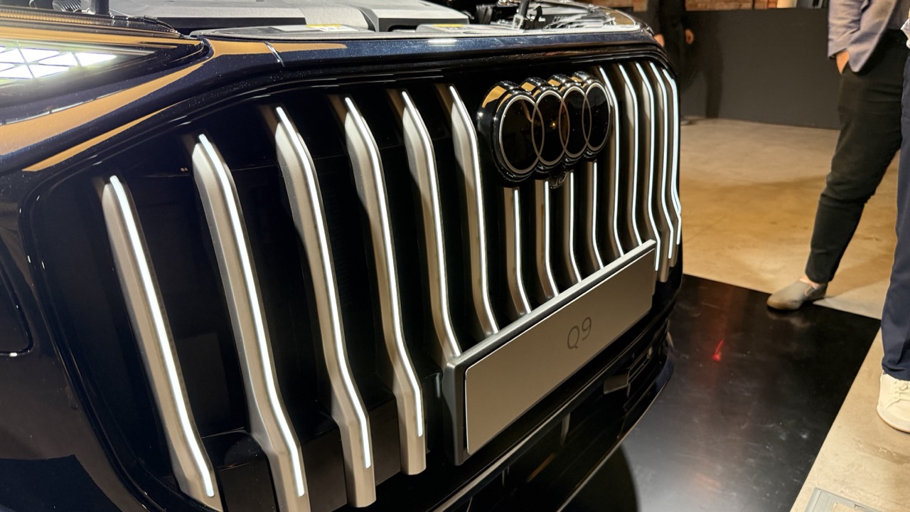 Zbliżenie na grill i logo Audi Q9, po prawej fragment nóg osoby.