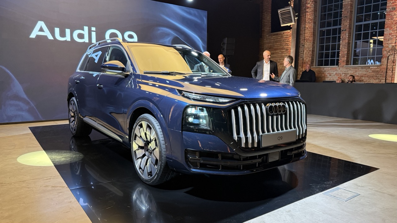 Niebieskie Audi Q9 na platformie, a w tle czterech mężczyzn przy ceglanych ścianach i oknach.