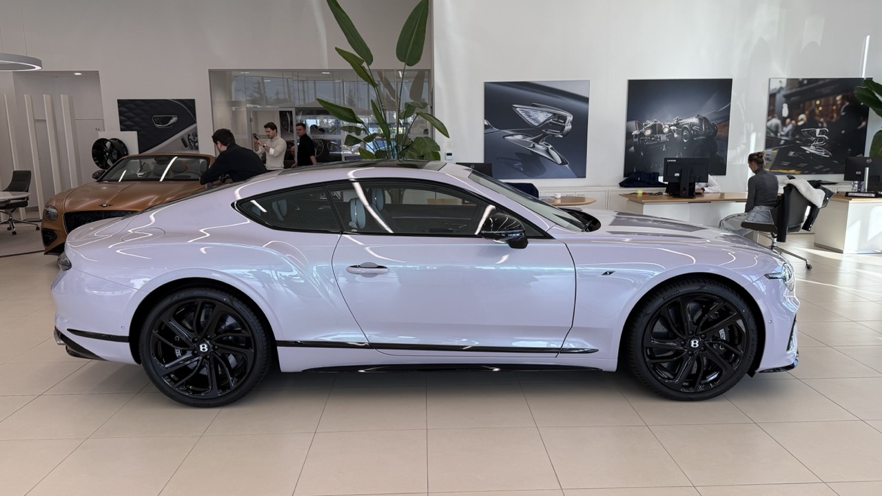 Jasnofioletowe coupe marki Bentley z czarnymi felgami w salonie samochodowym.