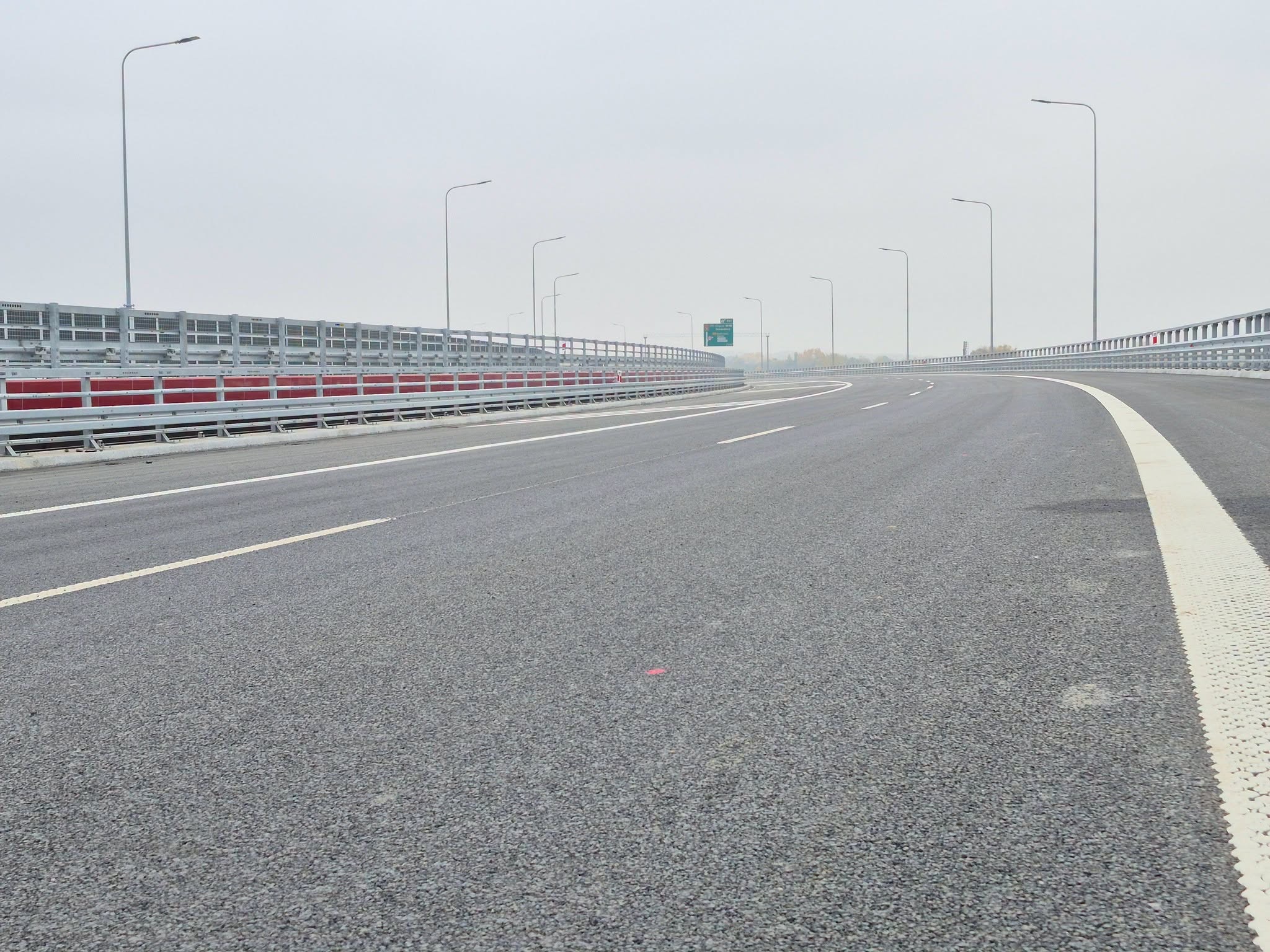 Kręta autostrada z asfaltowymi pasami ruchu i szarymi barierkami pod zachmurzonym niebem.