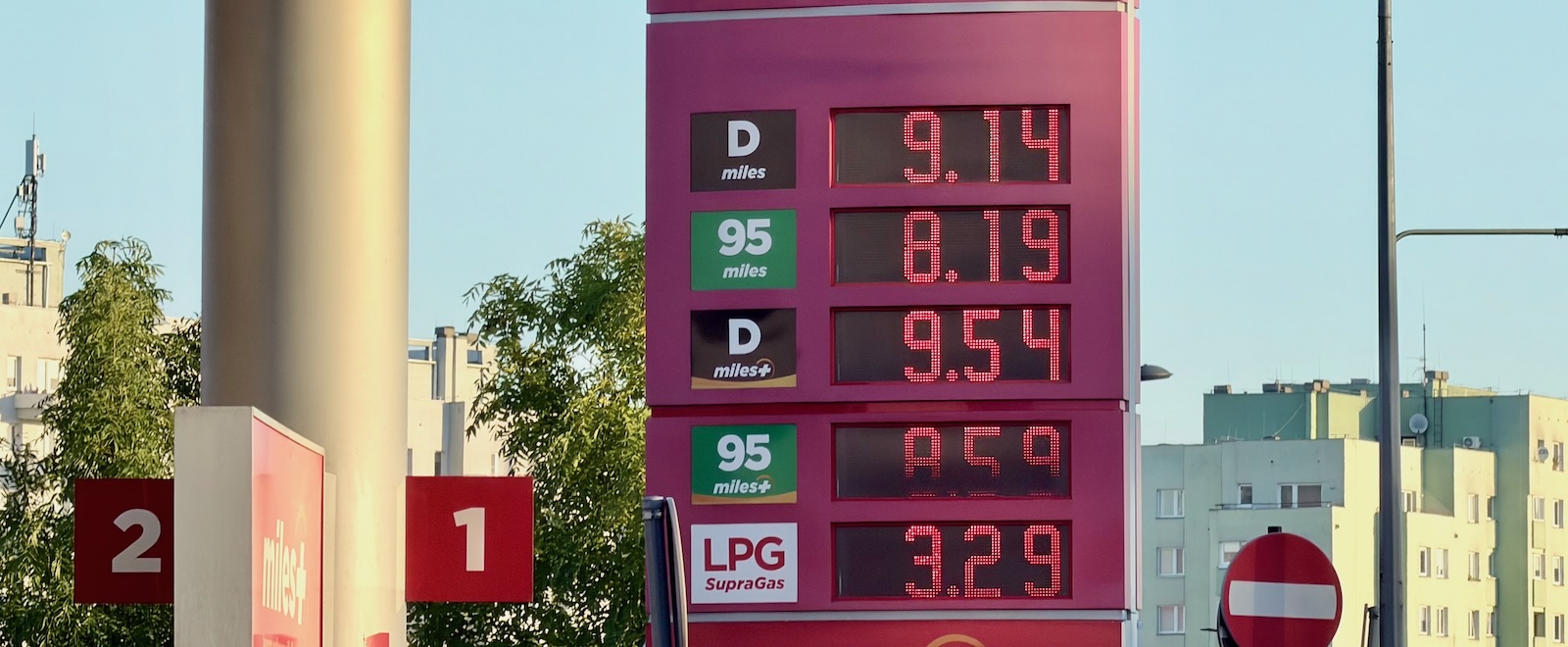Tabela cen paliw: 9,14; 8,19; 9,54; 8,59; LPG 3,29; w tle budynki i drzewa.