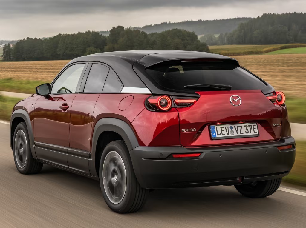 Czerwona Mazda MX-30 jedzie wiejską drogą wśród pól i drzew.