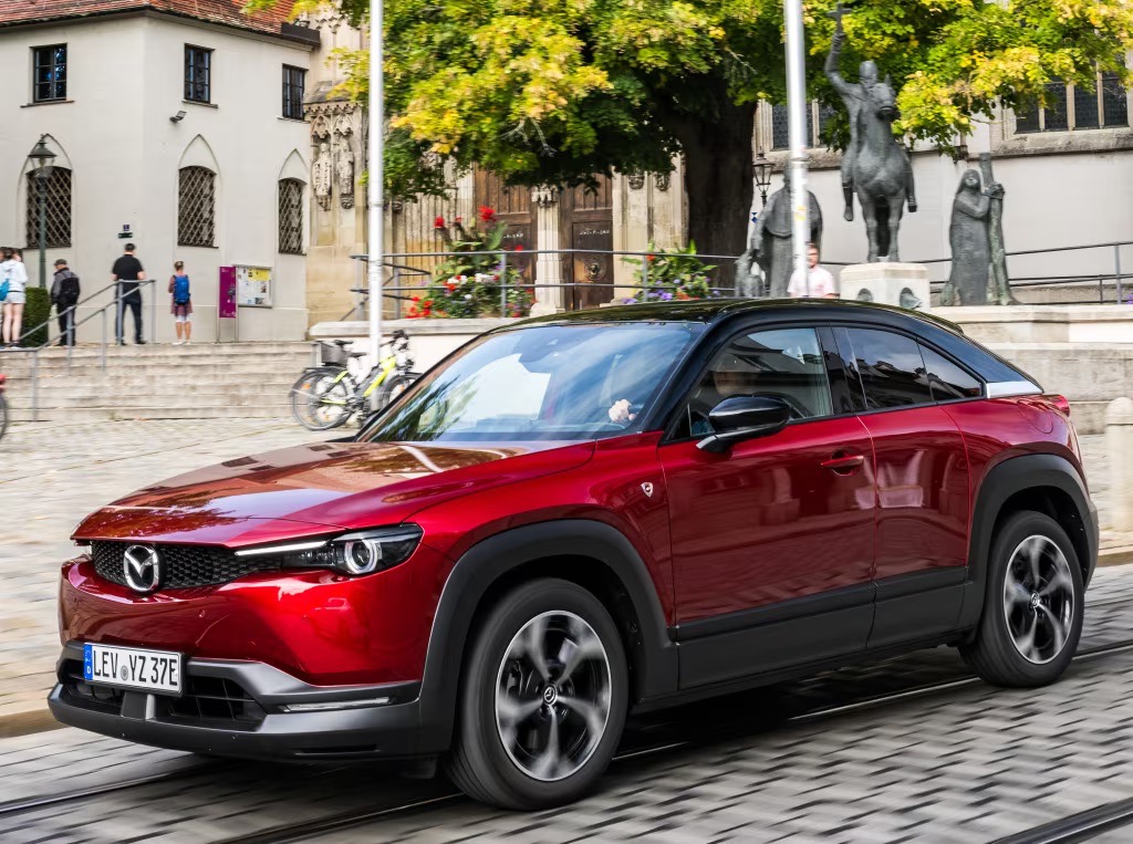 Czerwona Mazda MX-30 jedzie po brukowanej ulicy na tle zabytkowych budynków i pomnika.