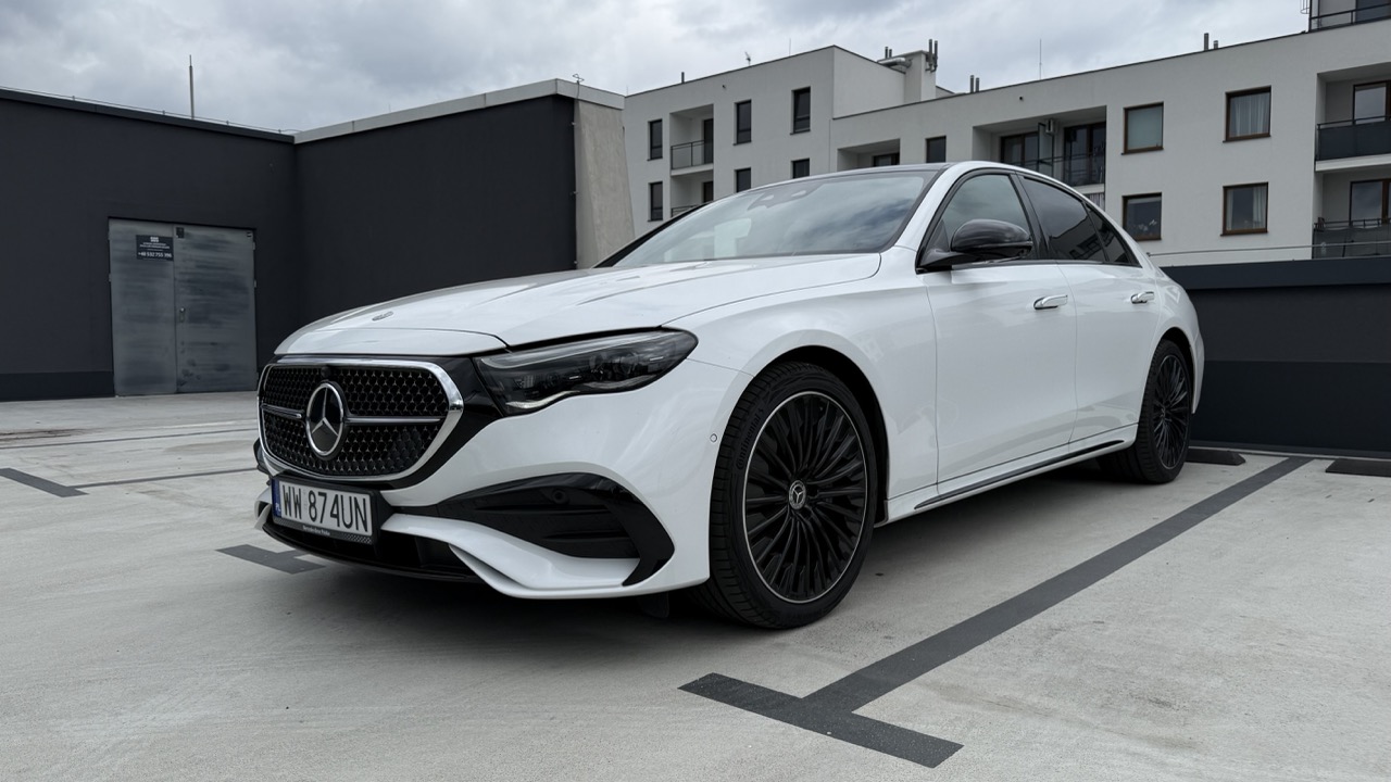 Biały sedan marki Mercedes-Benz zaparkowany na utwardzonym parkingu.