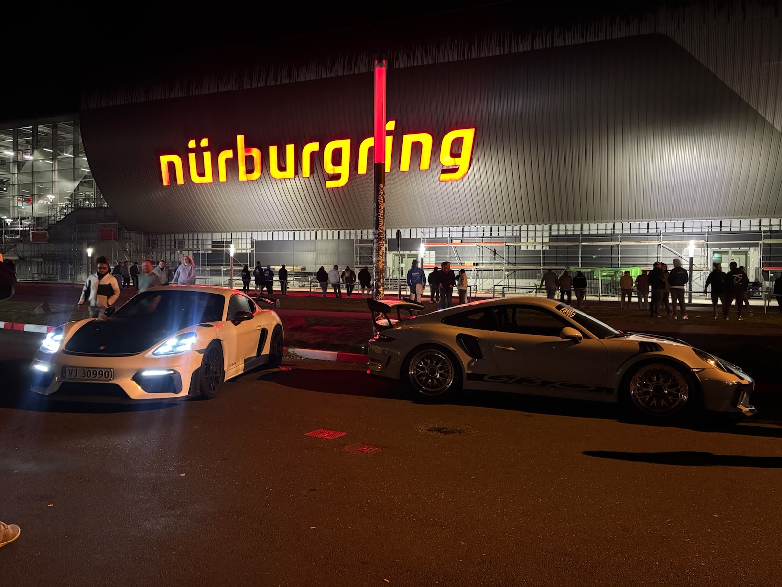 Dwa sportowe Porsche zaparkowane nocą przed wjazdem na tor Nürburgring.