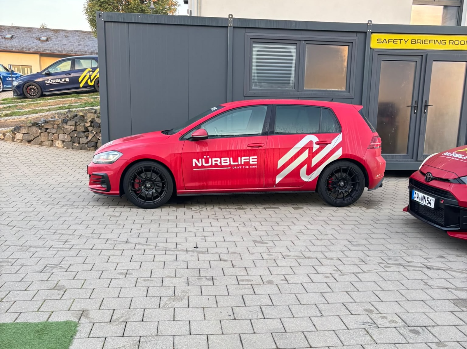 Czerwony Volkswagen Golf z logo Nürblife zaparkowany na utwardzonym parkingu toru Nürburgring.