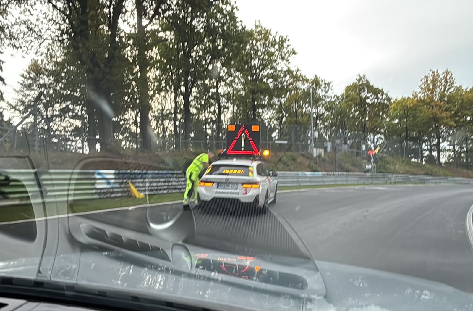 Ochroniarz w neonowej kamizelce stoi przy białym samochodzie ratunkowym toru Nürburgring z migającym znakiem.