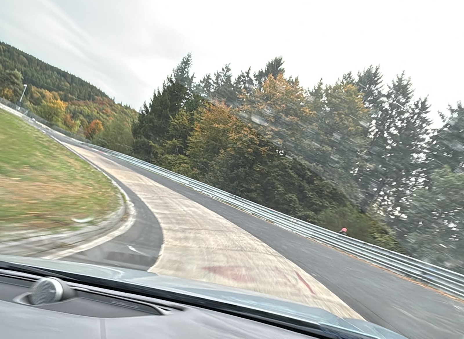 Widok z samochodu na zakręt toru Nürburgring otoczony drzewami.
