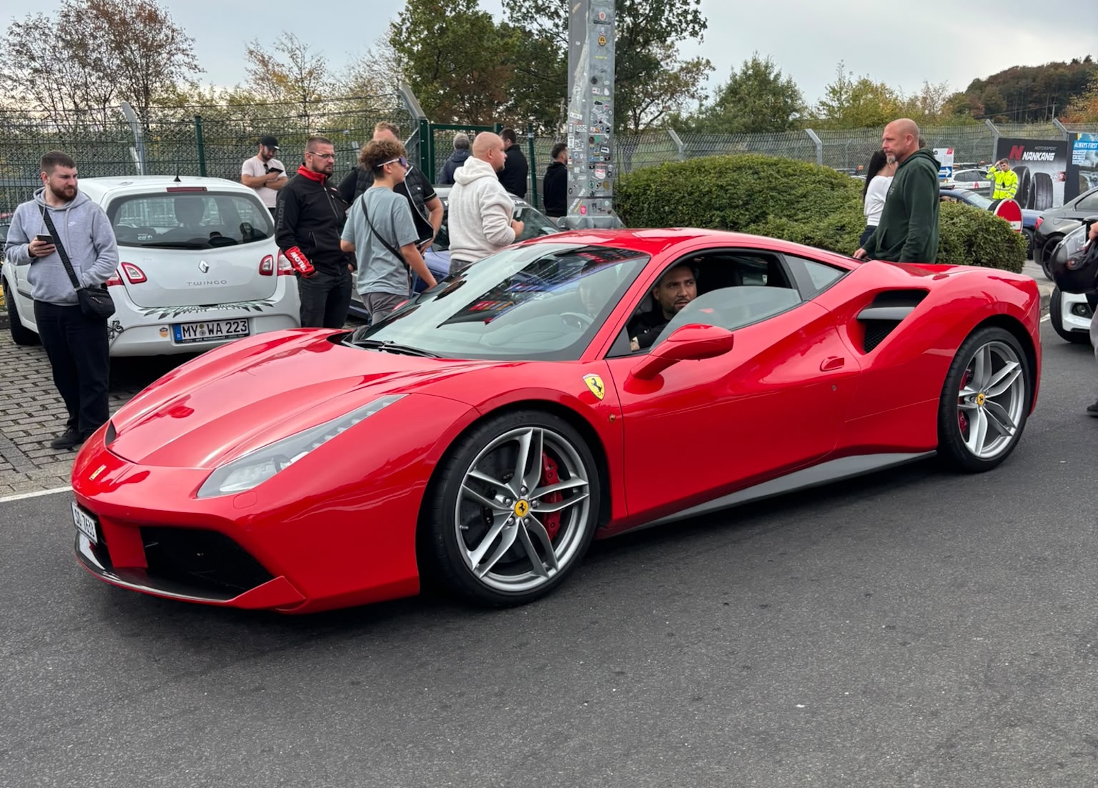 Czerwony samochód sportowy marki Ferrari z kierowcą w otoczeniu ludzi na parkingu przy torze Nürburgring.