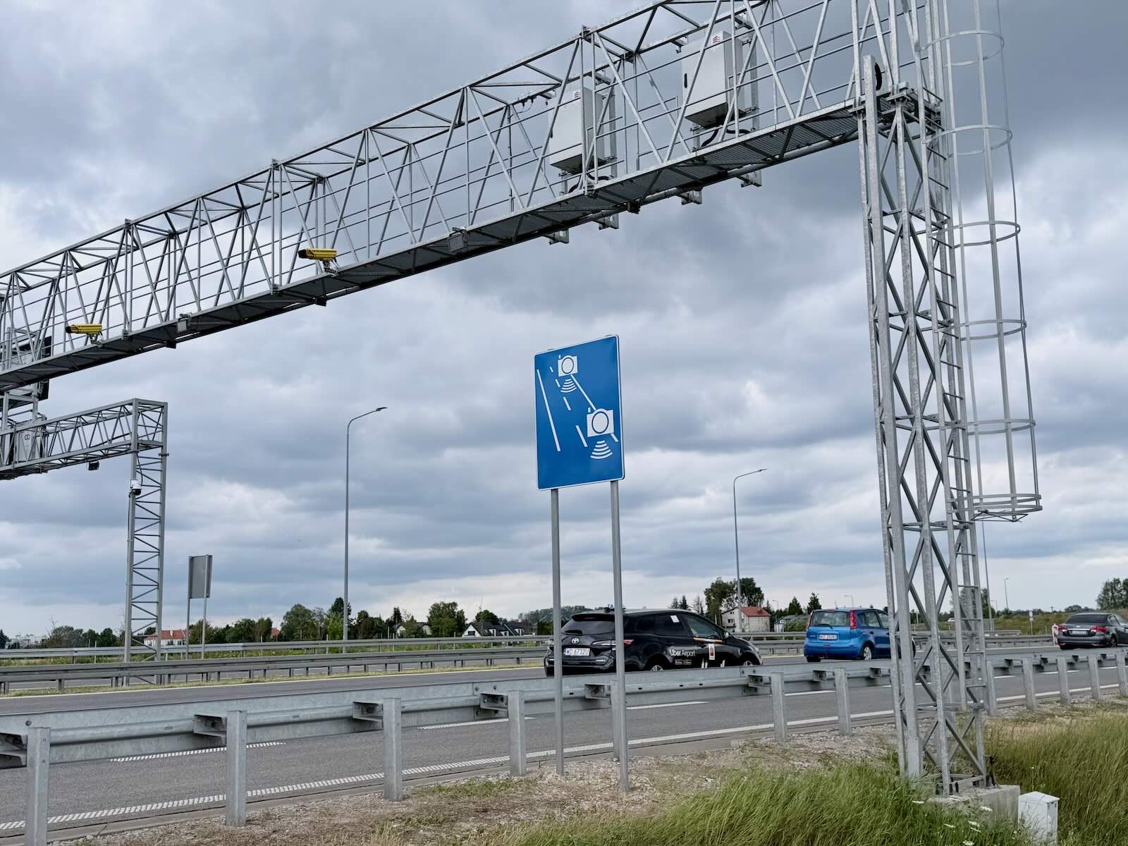 Niebieski znak drogowy informujący o fotoradarach i opłatach elektronicznych nad autostradą, po której przejeżdżają samochody.