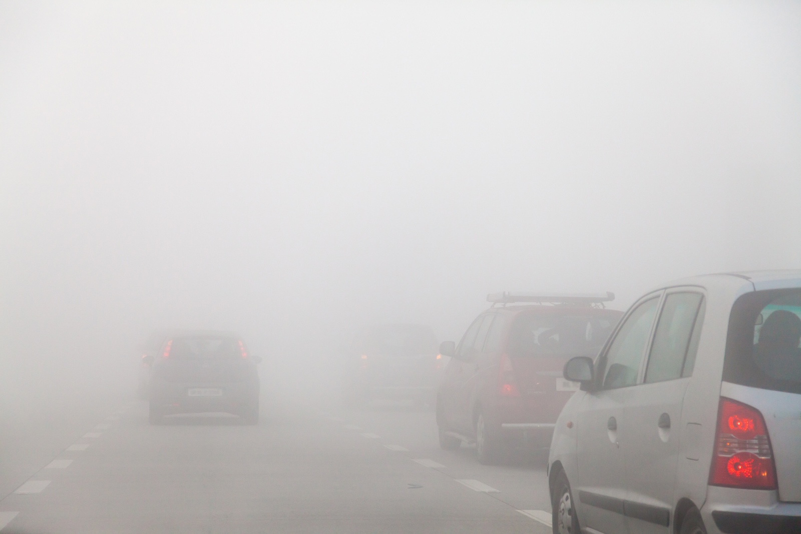Kilka samochodów jedzie autostradą we mgle, widoczność jest słaba, światła są włączone.